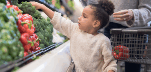 Photo of a toddler picking out vegetables during a trip to the grocery store with her parents.