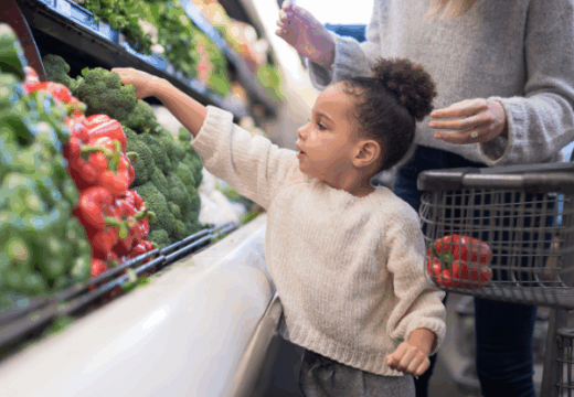 Photo of a toddler picking out vegetables during a trip to the grocery store with her parents.